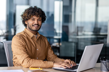 Portrait of young successful Indian man at workplace inside office, businessman smiling and looking at camera, man at work using laptop, programmer with curly hair coding software.