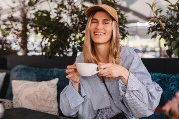 Wall Mural - Beautiful young woman looking at camera with coffee sitting in cafe and look happy. Blonde wearing stripped shirt and beige cap sitting on the sofa in restaurant. Coffee break.