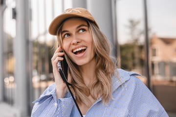 Wall Mural - Closeup portrait of a cheerful young woman making a phone call outdoors in the city walking near office. Happy woman wear stripped shirt and beige cap. Female look at side.