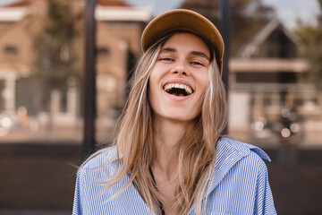 Wall Mural - Cute young woman with a lovely sense of humor standing leaning against a glass window exterior with copy space in an urban street laughing at the camera. Attractive blonde girl wear shirt and cap.