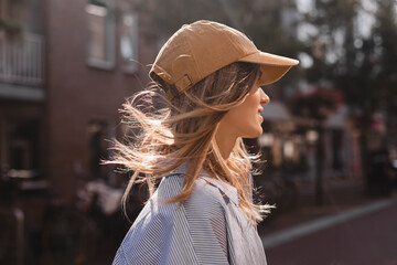 Wall Mural - Close up photo of young blonde woman walking outdoor on the street in city, wear cap, stripped shirt, flying hair. Woman look happy and smiling.