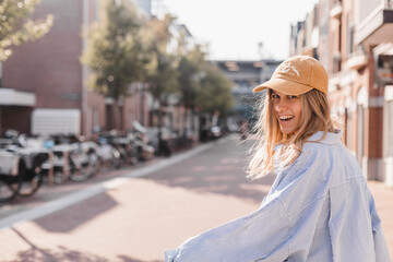 Wall Mural - Close up photo of young blonde woman walking outdoor on the street in city, wear cap, stripped shirt, flying hair. Woman look happy and smiling.