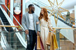 © Roman - bearded afro man in black pants, T-shirt and blue shirt looking at his ginger girlfriend in stylish white dress while walking at shop center, love, relationship