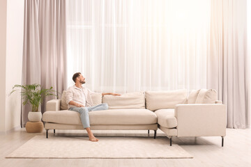 Wall Mural - Happy man resting on sofa near window with beautiful curtains in living room