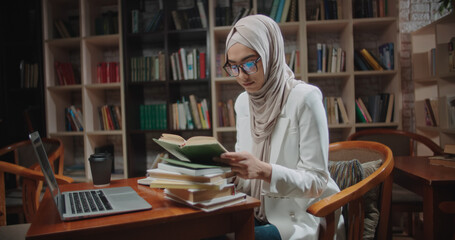 Wall Mural - Female muslim student is studying at desk in library, using laptop and books. Girl wearing hijab is preparing for exams - student lifestyle, modern Islam concept close up