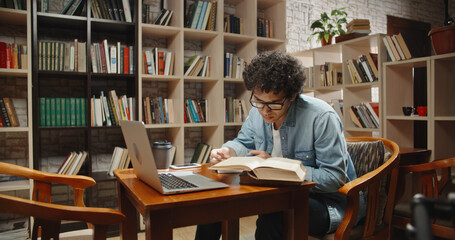 Asian student doing research in library using laptop in books, making a project and preparing for exam. Nerdy guy studying his lessons - education, self-study concept close up