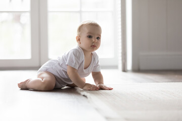 Cute little few month baby in white bodysuit learning to crawl on clean heating floor, sitting with open legs, looking away, touching carpet. Home portrait of adorable infant child