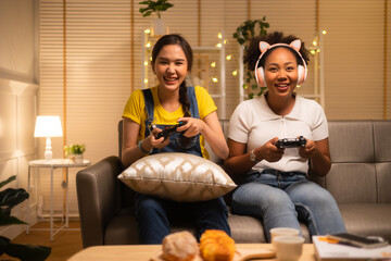 Wall Mural - Young  asian and african woman playing video games at home living room at night.Lesbian couples or lgbt activities on vacation inside the room.