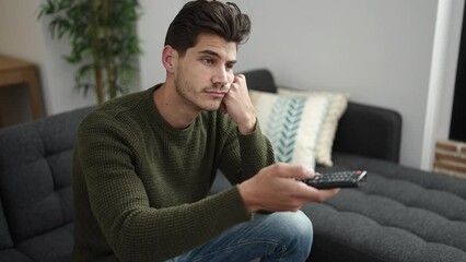 Wall Mural - Young hispanic man watching tv sitting on sofa with boring expression at home