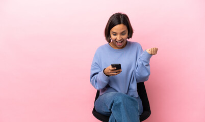 Young mixed race woman sitting on a chair isolated on pink background surprised and sending a message