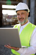© sakoqwakestock - Portrait of the builder in helmet and vest using wireless laptop for improving blueprints while standing at construction site. Concentrated bearded maleplanning system of building structures.