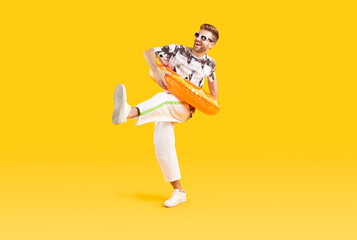 Wall Mural - Funny man enjoying his summer holidays at the beach. Happy, smiling guy in summer clothes, sunglasses and an inflatable swim ring walks isolated on a yellow orange color background. Full body portrait