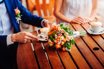 On the wooden table is the bridal bouquet and coffee mugs on a platter. Groom moves cup of coffee. The bride was hands on the edge of the table.