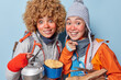 © WHstudio Leushin N - Happy cheerful active female campers look gladfully at camera going to have lunch in forest dressed in windbreaker have red frozen face stand closely to each other isolated over blue background