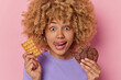 Headshot of curly haired young European woman holds tasty waffle and chocolate cookie licks lips wants to eat sweet appetizing food dressed in casual purple t shirt isolated over pink background