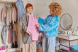 Stylish redhead woman chooses clothes from wardrobe going to put on pink jacket her friend helps with choice of outfit pose in bedroom. Two female models get dressed on party. Fashion concept