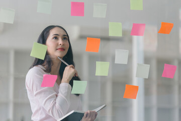 A professional millennial Asian businesswoman writing on colorful sticky notes and attaching them to a glass window in a creative office. Business planning and brainstorming strategy concept.