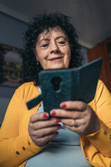 Front vertical image of an older woman sitting in bed looking at her mobile phone with book cover. Selective focus on the woman.