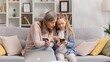 © YURII MASLAK - Young granddaughter teaches her senior grandmother to use a card for shopping on the Internet. A 60s grandmother and a little blond granddaughter are buying something by phone payment.