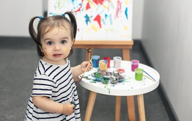 A 1.5-year-old girl with two tails in a striped dress paints with paints at an easel. There is a table with paints and brushes nearby. The girl looks into the frame. Child perception, children's creat