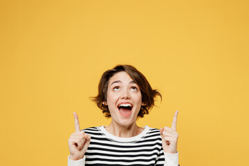 Young excited fun woman wear casual striped black and white shirt point index finger overhead indicate on workspace area copy space mock up isolated on plain yellow color background studio portrait.