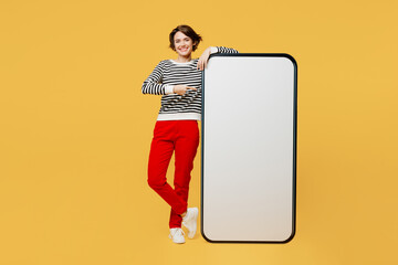 Full body young woman wears casual black and white shirt point index finger on big huge blank screen mobile cell phone smartphone with workspace mockup area isolated on plain yellow color background.