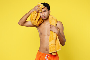 Exhausted sad tired sick ill hot young sexy man wear orange shorts swimsuit wiping sweat with towel relax near hotel pool isolated on plain yellow background. Summer vacation sea rest sun tan concept.