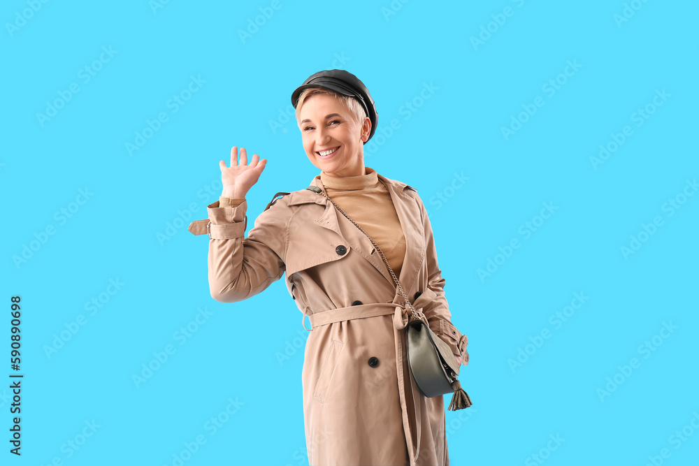 Stylish mature woman waving hand on light blue background