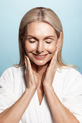 Wall Mural - Aging beauty. Vertical portrait of middle aged, elderly woman, 50 years, touches her smooth, nourished skin, applies facial cream, moisturizer, smiling pleased, wears bathrobe, blue background