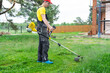 © Ольга Симонова - Male gardener mows the green grass of the lawn in the backyard at construction site with a gasoline mower. Trimmer for the care of a garden plot
