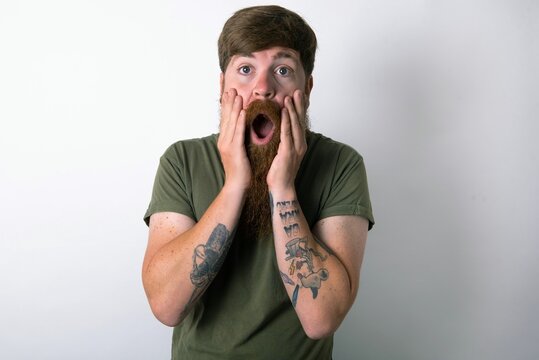 Scared terrified red haired man wearing green T-shirt over white studio background shocked with prices at shop, People and human emotions concept