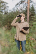 © junky_jess - A woman park ranger in uniform looks through binoculars at the forest area with a clipboard in hand in summer, vertical photo, selective focus.