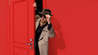Stylish bearded man in trench coat and glasses, detective peeking out red door and attentively looking. Finding out secrets. Concept of emotions, facial expression, lifestyle