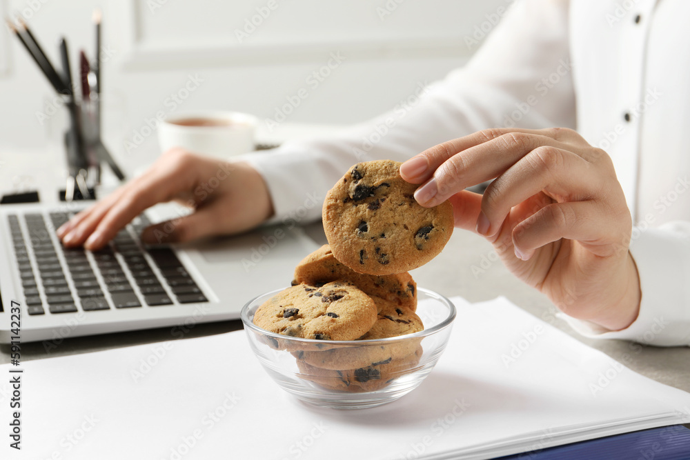 Woman taking chocolate chip cookie from bowl while working with laptop ...