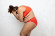 © Юля Бурмистрова - Naked overweight woman bend over holding head by hands side view, white background. Woman in red underwear with cellulite hips and large hanging belly. Suffer from pain, fat figure, plus size people.