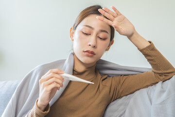 Wall Mural - Sick, influenza asian young woman, girl headache have fever, hand touching forehead, holding thermometer for check measure body temperature, illness while sitting rest on sofa at home. Health care.