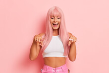 Wonderful girl with beautiful long hair stands on a pink background, wears a white top and pink pants, points fingers down at your advertisement, looks down and smiles happily, copy space, high