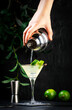 Gimlet alcoholic cocktail with dry gin, liqueur, lime juice and ice in glass garnished with lime zest. Bartender pours drink from shaker into glass. Black background