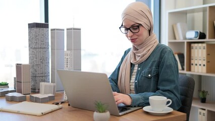 Joyful muslim female wearing hijab and glasses typing on laptop while holding architectural model from office desk indoors. Proficient urban planner developing land use programs of metropolitan area.