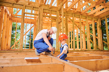 Father With Toddler Son Building Wooden Frame House. Builder Teaching His Son How To Use Screwdriver On Construction Site, Wearing Helmets And Blue Overalls On Sunny Day. Carpentry And Family Concept.