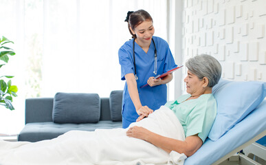 Asian professional successful female internship nurse in blue uniform with stethoscope visiting old senior elderly pensioner woman patient laying lying down on bed writing symptoms down on clipboard