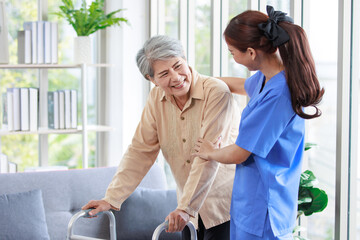 Asian young professional successful friendly female nurse in blue hospital uniform helping supporting physical therapy senior old pensioner unhealthy injury woman patient walking via four legs walker