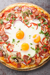 Freshly baked pizza with bacon, eggs, peppers, mushrooms, mozzarella and red onions close-up on a wooden board on the table. Vertical top view from above