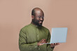 © pressmaster - Smiling African American man holding laptop computer and typing on keyboard while standing on brown background