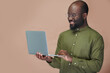 © pressmaster - Smiling African American man in eyeglasses working online using his laptop standing on brown background