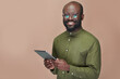 © pressmaster - Portrait of African American man in eyeglasses smiling at camera while using tablet pc against brown background