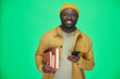 © pressmaster - Portrait of African American man with books smiling at camera while using his smartphone against green background