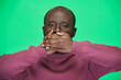 © pressmaster - Portrait of African American man looking at camera closing his mouth with hands against green background