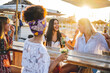 © MandriaPix - four women having fun at the beach bar, young female friends laughing and chatting, having some drinks and spend time on vacation, holiday and summer concept