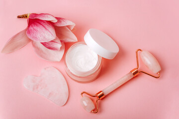 Wall Mural - Rose quartz crystal facial roller and gua sha scraper, face cream, magnolia flower on pink background. Facial massage kit for lifting therapy. Top view, flat lay